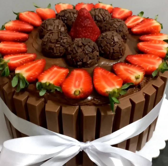 Bolo decorado com barras de chocolate, morangos fatiados e brigadeiros no topo, com laço branco