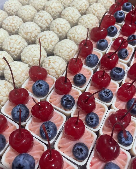 Brigadeiros de chocolate branco ao lado de copinhos com creme, cereja e mirtilo