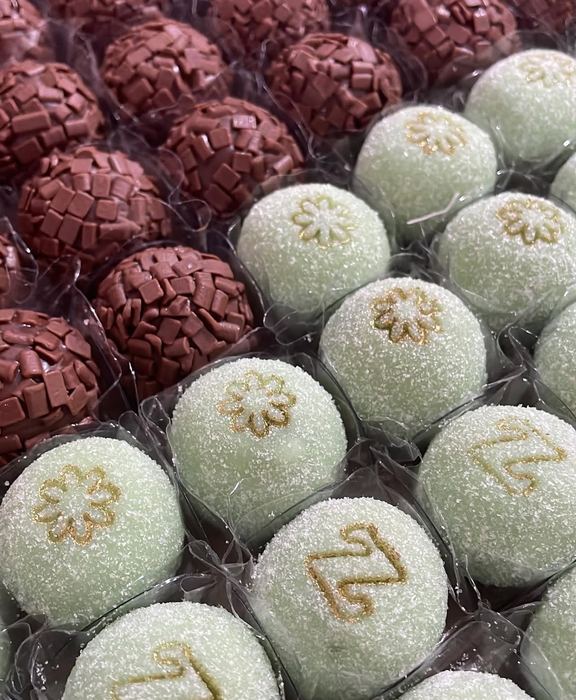 Brigadeiros de chocolate ao lado de doces verde menta com inicial e flor douradas aplicadas à mão