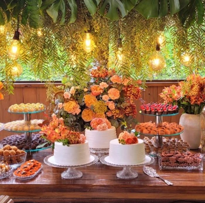 Mesa de doces completa montada com três bolos brancos, arranjos florais e bandejas de doces finos em festa na Cidade de Curitiba